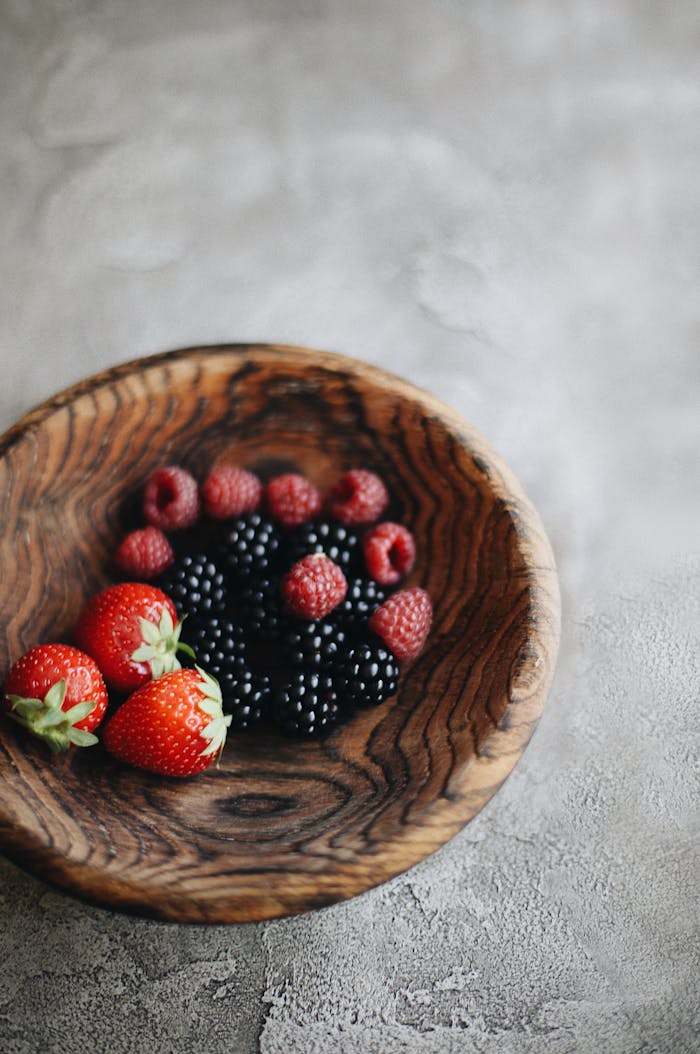 A combination of strawberries, blackberries, and raspberries in a wooden bowl on a textured surface.